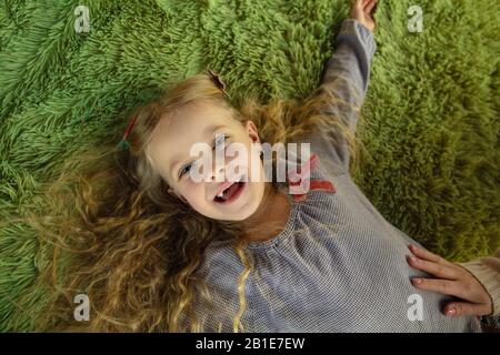 Happy cute caucasian girl, daughter laying down and laughting. Playing, having fun and looks happy. Lovely curly blonde kid. Mother's day, celebration, weekend, holiday and childhood concept. Stock Photo