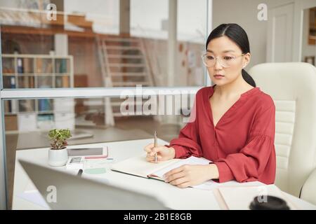 Portrait of modern Asian businesswoman wearing red blouse working at desk in office cubicle, copy space Stock Photo