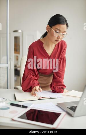 Portrait of successful Asian woman leaning over desk while working in white office interior Stock Photo