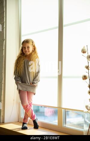 Happy cute caucasian girl, daughter posing against window and smiling. Playing, having fun and looks happy. Lovely curly blonde kid. Mother's day, celebration, weekend, holiday and childhood concept. Stock Photo