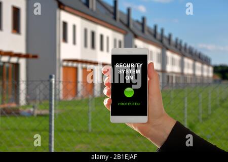 Woman holding a smartphone with home security system panel on blurred house background Stock Photo