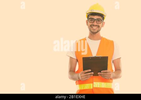 Studio shot of young happy Persian man construction worker smiling while holding clipboard Stock Photo