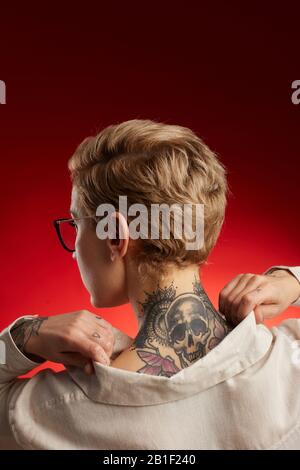 Back view of short haired young woman with neck tattoo featuring scull and butterflies posing against red background in studio Stock Photo