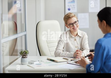 Portrait of smiling young woman talking to client or partner while working at desk in office cubicle, copy space Stock Photo