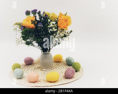 Spring flower arrangement with yellow roses and purple blossoms one a cream colored round placemat surrounded by string decorated Easter eggs in yello Stock Photo