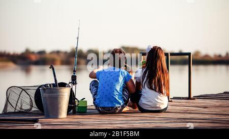 Two young cute little friends, boy and girl eating sandwiches and fishing on a lake in a sunny summer day. Kids are playing. Friendship. Stock Photo