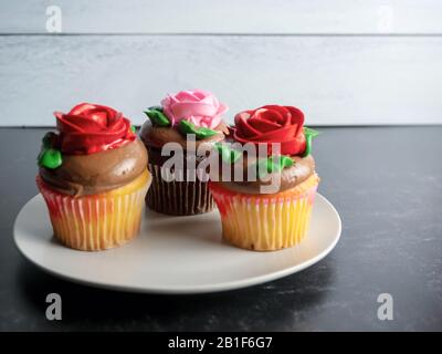 3 cupcakes sitting on a plate, yellow and chocolate with chocolate frosting and red and pink roses made from frosting, on a black slate surface with a Stock Photo