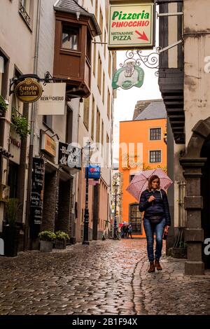 A woman wears a pink umbrella to protect herself from the rain. She walks on the wet cobblestones of an historical alley in Cologne. Stock Photo