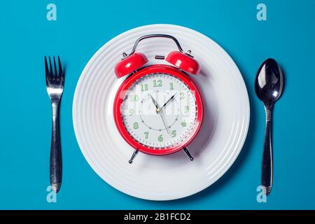 Red alarm clock on the white plate with spoon and fork on the color background. Food and diet concept
