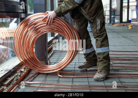 Male plumber worker in green uniform holds large round skein of new shiny copper pipes, lay pipeline, installation tubes on floor with warm heating  o Stock Photo