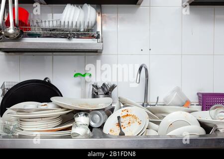 Large metal sink with dirty dishes in professional restaurant kitchen, stack of unclean white plates, crockery, appliances with leftover food, water t Stock Photo