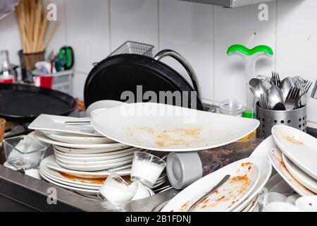 Large metal sink with dirty dishes in professional restaurant kitchen, heap of unclean white plates, crockery, appliances with leftover food, water ta Stock Photo