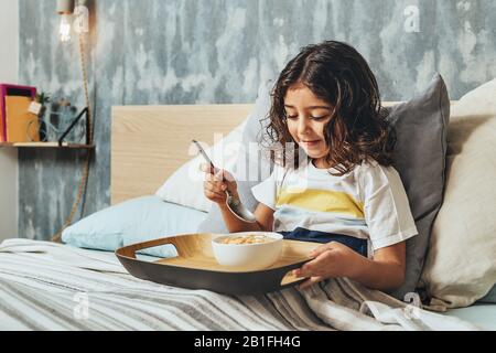 little girl having sunday breakfast in her parents' bed. Kid enjoying a bowl of cereal at home, concept of healthy living and eating Stock Photo