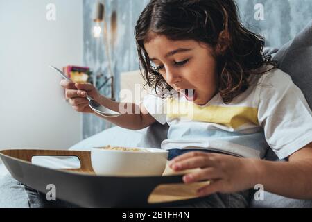child surprised while having sunday breakfast in her parents' bed. Little girl enjoying a bowl of cereals at home in the morning, concept of healthy l Stock Photo