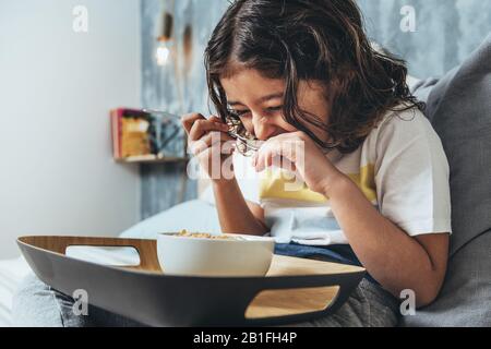child laughing while having sunday breakfast in her parents' bed. Little girl enjoying a bowl of cereals at home in the morning, concept of healthy li Stock Photo