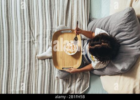top view of a little girl having sunday breakfast in her parents' bed. Kid enjoying a bowl of cereal at home, concept of healthy living and food Stock Photo
