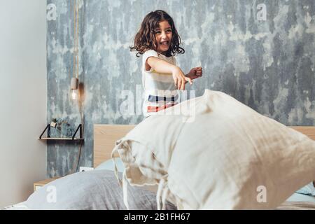 funny little girl standing on her parents bed smiling and throwing a cushion in a pillow fight, home lifestyle concept and happy childhood Stock Photo