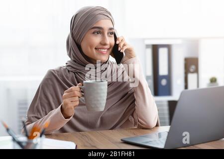 Muslim female manager talking on cellphone and drinking coffee at workplace Stock Photo
