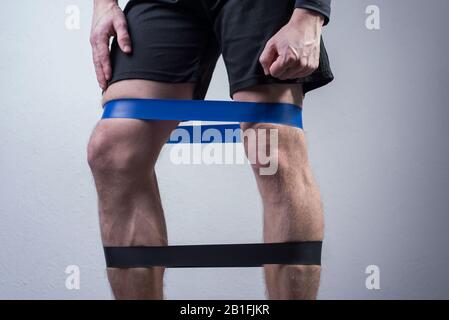 closeup, bottom view of the guys muscular legs in sports shorts, stretching fitness elastic bands with his legs, involvement of leg muscle groups that Stock Photo