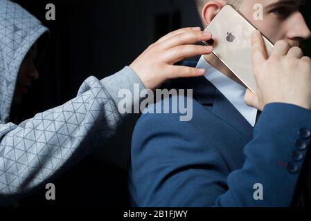 Zaporozhye. Ukraine. February 2020. Close-up portrait of a young bearded guy in a business suit holding an iPhone XS max. A robber in a dark hood, try Stock Photo