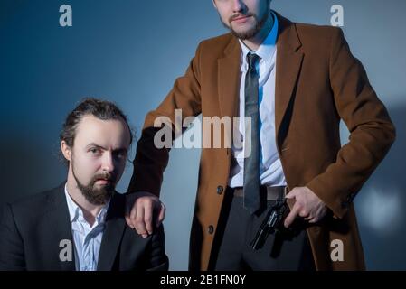 Portrait of two men in business suits, one of them is sitting, the other is holding a gun. On a gray background. Dramatic light. Artistic setting. The Stock Photo