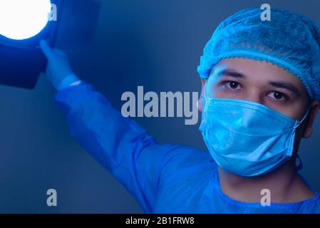 close-up portrait of a caucasian doctor surgeon, in a sterile suit, mask, holds a lamp, pointing it to the camera. On a blue background. Operating roo Stock Photo