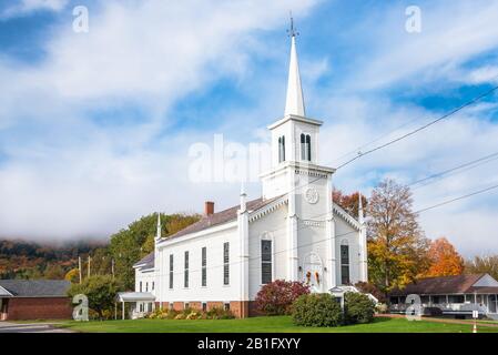 Traditional white American wooden church at the foot of a forested hill shrouded in morning fog on a autumn day