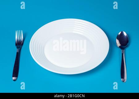 White plate, fork and spoon on blue color background. Stock Photo
