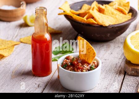 Two bowls full of salsa dip, hot red chili sauce bottle and tortilla chips on rustic wooden table, sause being added to one of the bowls Stock Photo
