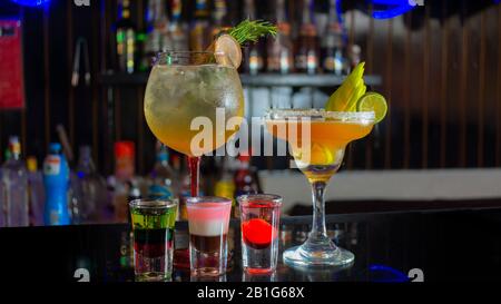 Glass cup with passion fruit margarita accompanied by three small glass glasses with tequila reflecting on the table of a bar with unfocused backgroun