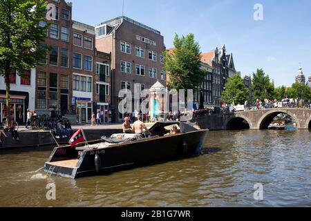 View of people riding a open boat on canal doing a cruise tour in Amsterdam. Many people hang out on street and crossing Armbrug bridge. It is a sunny Stock Photo