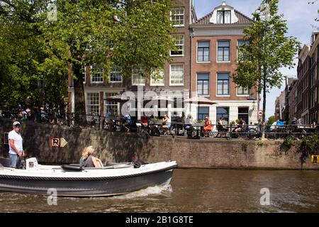 View of people hanging out by canal at a cafe and riding a small open boat, trees, historical and traditional buildings showing Dutch architecture sty Stock Photo