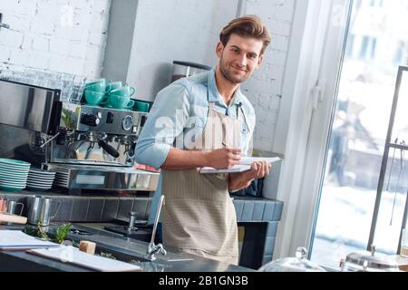 Coffee Passion. Young barista at coffee shop taking noted on new recipe smiling joyful Stock Photo
