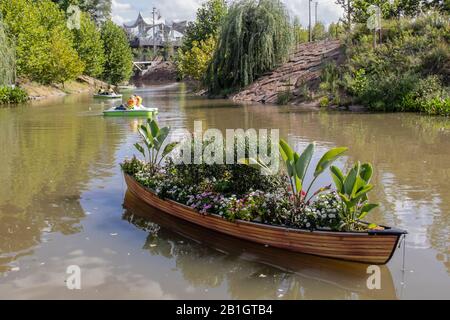 8-18-2018 Tulsa USA Vintage wooden canoe filled with plants floats in river at Gathering Place public park with people in paddle boats and Boathouse i Stock Photo