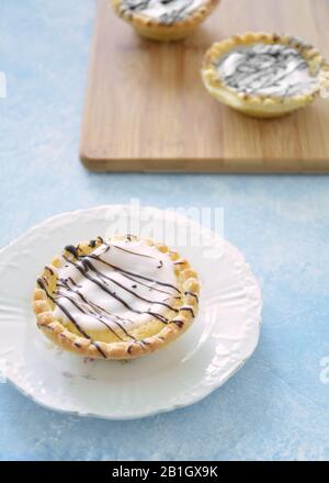 Iced almond tarts with chocolate drizzle on bamboo board and white plate in vertical format. Light blue background.  Selective focus on front tart. Stock Photo