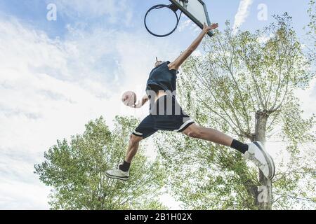 Basketball player performing huge slam dunk on the court in urban grunge camp with back light - Yong athlete in action outdoor - Sport concept - Origi Stock Photo