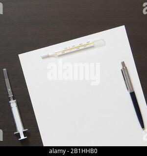 Template of white paper with pen, thermometer and syringe for injection on dark wenge color wooden background. The provision of medical and pharmacological services. With empty space for text. Stock Photo