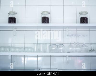 White cafe decoration minimal style. Coffee beans in three glass bottle, various style of glass on white wooden shelf on white mosaic grid wall backgr Stock Photo