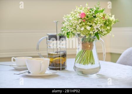 Table with white tablecloth tea in teapot, cups and bouquet of beautiful flowers Stock Photo