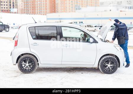 Novosibirsk, Russia - 02.17.2020: A man stands near a broken car with an open hood and looks at the engine to identify a breakdown while waiting for r Stock Photo