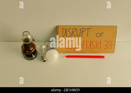 Closeup and focus on cardboard sign with handwriting DISRUPT or BE DISRUPTED placed on table among candle, oil lamp, incandescent lamp and LED lamp Stock Photo