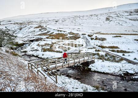 A walker crosses a bridge over a stream on the footpath towards Pen y Fan mountain on Brecon Beacons National Park, Wales, after temperatures plummeted overnight across Britain, and forecasters warned of more ice and snow over the next 24 hours. Stock Photo