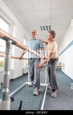 Physiotherapist helping senior man in rehabilitation walking on the bars Stock Photo