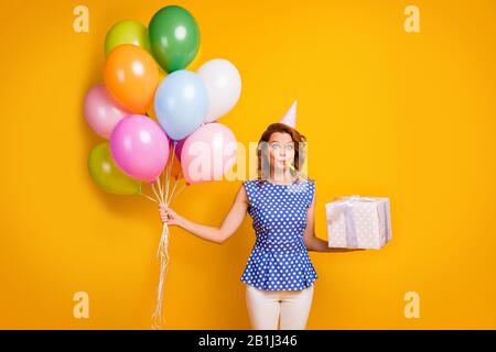 Photo of pretty lady hold many colorful air balloons big giftbox party blow noise maker wear paper cone cap polka-dot blue shirt white trousers Stock Photo