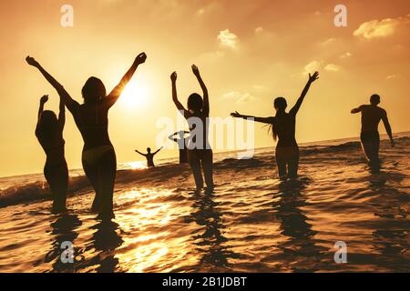 Big group of happy peoples stands at sunset sea beach. Silhouettes with raised arms