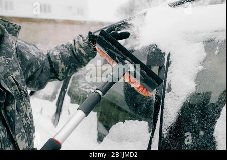 man in camouflage clothing without a face cleans the car from heavy snow with a black brush with a red pile. Cleaning the snow-covered glass of the ca Stock Photo