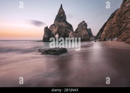 Rocky sea stacks at Portugal Ursa Beach at Atlantic Ocean coast in sunset light. Foamy waves rolling on picturesque landscape vacation scene Stock Photo