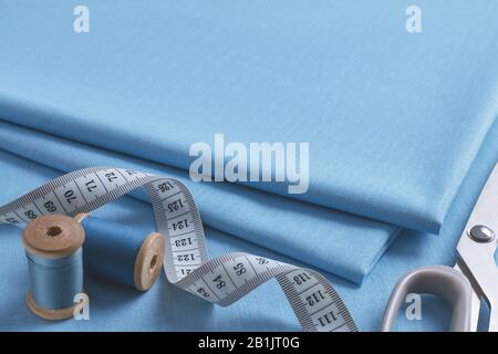 Set of tailoring accessories. Close up of a stack of folded blue fabric, sewing scissors, a twisted white measuring tape and two spools of thread Stock Photo