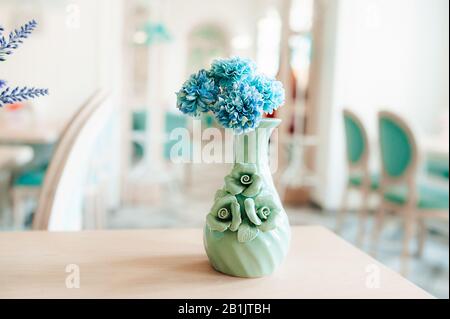 A small mint colored vase with blue cornflowers on a wooden table in a bright spacious room with blue chairs Stock Photo
