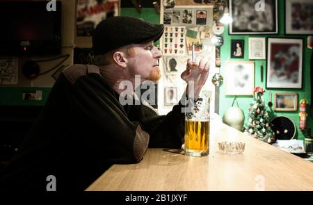 Red haired almost Irish guy seating in the bar and smokes cigarette and drinks some beer Stock Photo